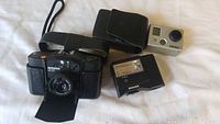 Minox 35 GT black film camera with leather case and strap, Minox FC 35 black flash unit, GoPro HERO2 silver digital camera with protective housing, laid out on white cloth surface.