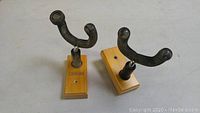 Close-up image showing two used guitar hooks with black metal arms mounted on rectangular wooden blocks, placed on a white surface.