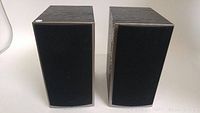 Pair of ATHENA bookshelf speakers with black fabric grille cover installed, woodgrain finish visible on sides and top.
