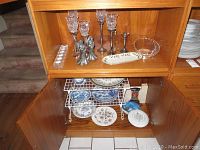 Cabinet shelf showing multiple angel-shaped metal candle holders with glass globes of varying heights. Below, multiple ceramic plates and bowls with Asian and English floral patterns.