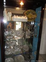 Glass cabinet shelf with white Last Supper ceramic sculpture, two white shepherd figurines on left, green ceramic grapevine plate on right, and assorted clear glass and crystal bowls below.