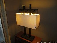 Photo shows one of the two matching lamps lit, featuring rectangular beige fabric shade and heavy metal upright frame on rectangular base.