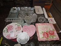 Overview of assorted glass and ceramic serving ware on a table, showing floral patterned ceramic plates, clear textured glass platters and bowls, and a pink heart-shaped plate with 'LOVE' text.