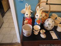 Photo showing the basket with marble-like eggs in pastel colors, two resin rabbit figurines, and other small decorative eggs and containers on a dark surface