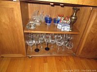 Shelves inside wooden cabinet showing assorted glassware, glass birds, porcelain pieces, metal vase, and coasters.