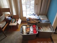 View of bins and cardboard box filled with assorted folded fabric items including blankets, throws, and table covers near a door.