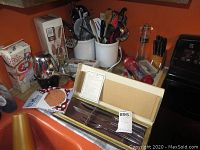 Overview photo of kitchen counter with cooking utensils in crocks, boxed Carvel Hall cutlery set, ice crusher, and other items