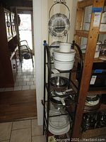 Full view of metal rack holding mixing bowls, colanders, and ceramic bakeware, with corner wall plates displayed on hanger.