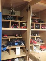 Assorted vintage model vehicles displayed on wooden shelves in a glass-door cabinet, including cars with red spoked wheels and a larger blue vehicle.