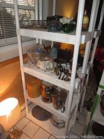 Four white shelving unit with mixed items including silver plated trays, metal containers, glass, and ceramics