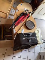Photo of 3 umbrellas, wooden frame wall clock, black hard case luggage, and part of cardboard box for picture frames and towels.