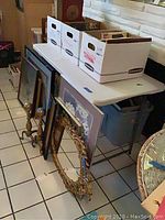 White folding collapsible table with boxes on top, leaning picture frames and table fan on the ground nearby