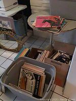 Boxes and bin full of vintage vinyl records, cassettes, and VHS tapes on floor near table and mirror