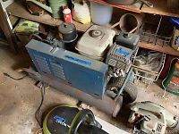 Side view of the blue Emglo gas powered air compressor with visible motor, wheels, hose, and attached components in a workshop environment.