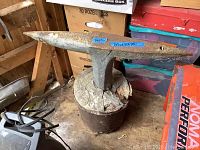Antique anvil on top of a metal bucket filled with cracked cement, showing wear and rust.