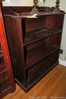 Wooden 4-shelf bookcase showing upper shelves and top raised backboard with wear and wear marks visible.