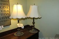 Pair of matching 26 inch table lamps with ornate bases and cream bell-shaped shades on a wooden surface against a wall with framed print.