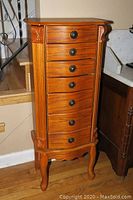Full front view of the tall wood jewelry chest showing six drawers with dark metal pulls, carved corner details, and cabriole legs.