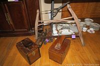 Photo showing two wooden shoe shine boxes in front and seven long rustic wooden wall-mounted candle holders arranged vertically behind them on the floor, with additional background of stacked dishes and wooden furniture.