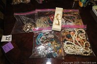 Overall assortment of necklaces and bracelets in multiple resealable plastic bags and one jewelry box on a dark surface.