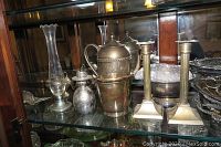 Silverplate teapot, metal bucket, creamer, sugar dish, clear glass vase, pair of candlesticks, metal dolphin bottle opener, and snowman figurine on glass shelf.