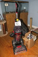 Full view of Bissell upright deep cleaner showing handle, water tanks, power cord, attached black bag, and nearby box with attachments.