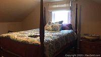 View of the queen size wooden poster bed with carved posts, made and set up with bedding in a bedroom with slanted ceiling and window in background.