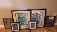 Group photo of seven framed wall art pieces leaning against a wall, showing different sizes and styles, mainly in dark brown frames with floral and architectural prints.