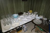 Wide view showing multiple glassware items, bowls, dishes, and plates arranged on folding tables in a basement setting with wood panel walls.