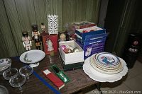 Overview photo showing nutcrackers, collectible Lenox plates and platters, assorted tree ornaments, glassware, and boxed holiday decorations on a table against wood paneling.