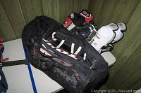 Black mesh outdoor carrying bag containing hockey pads, Reebok skates, and shin guards displayed on a table.