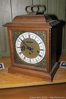 Front side view of Hamilton wooden table clock on a wooden surface showing the clock face with Roman numerals and ornate corner detailing.