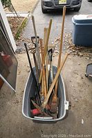 Plastic bin filled with various yard and construction tools with wooden handles visible in a garage doorway