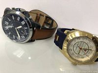 Photo showing both watches from an angled perspective, the Fossil watch has a black dial with Roman numerals and brown leather strap; the Hilfiger has a gold-tone bezel and blue strap.