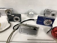 Overview of five digital cameras including Olympus, Sony, Kodak, and two others, arranged on white surface