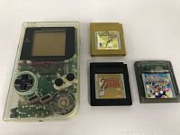 Front view of transparent Nintendo Game Boy with three game cartridges laid next to it: Pokemon Yellow, The Legend of Zelda DX, and Wario Land II.