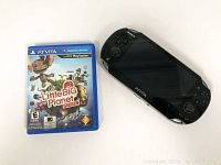 Front view of Sony PS Vita console next to LittleBigPlanet game case showing front cover art.