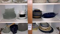 View of cabinet shelves showing clear glass plates and bowls on the left, white ceramic dishes with a wavy rim on the left top shelf, dark blue and blue-patterned stoneware on right and bottom right shelves.