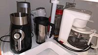 Front counter top view showing Hamilton Beach coffee grinder, travel mug, and white drip coffee maker with glass carafe near a sink.