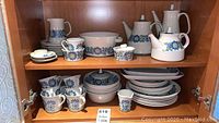 Two shelves of two incomplete vintage dishware sets with floral blue and green patterns including cups, saucers, plates, bowls, pitchers, teapots, and sugar bowls.