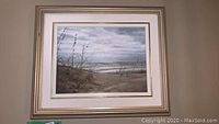 Framed oil painting showing sand dunes and sea oats with water and cloudy sky in background.