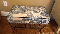 Photo showing the bench with upholstered cushion in toile fabric and curved black metal frame from the front.