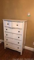 Front view of white wooden dresser showing 6 drawers with metal knobs against a tan wall and hardwood floor.