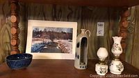 Photo showing 11x14 framed landscape painting, two ceramic oil lamps with floral decoration, a blue decorative bowl, and a ceramic vase with blue circular accents on wooden shelf