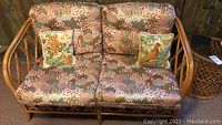 Front view of rattan love seat with floral cushions and two embroidered animal-themed pillows.