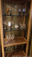 Glass cabinet with multiple glassware items on three shelves, including amber goblets, blue stemmed cocktail glasses, clear wine glasses, and a small glass bowl with shells.