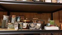 Full shelf view showing copper kettle, pitchers, mugs, Universal candleholder box, and other ceramic items.