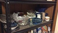 Shelf with a mix of kitchen items including blue divided fondue plates, ceramic coffee cup, metal cocktail shaker, glass bowls, and dark enamelled pots.