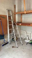 Photo showing both a 7 foot aluminum extension ladder leaning against the wall and a smaller aluminum step ladder standing open on the ground in a garage or storage area.