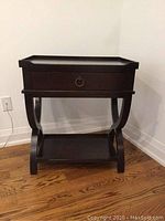 Front view of a dark-stained wooden end table showing single drawer with ring pull, curved legs forming X shape, and lower shelf. Light wear and scratches visible on wood surface.